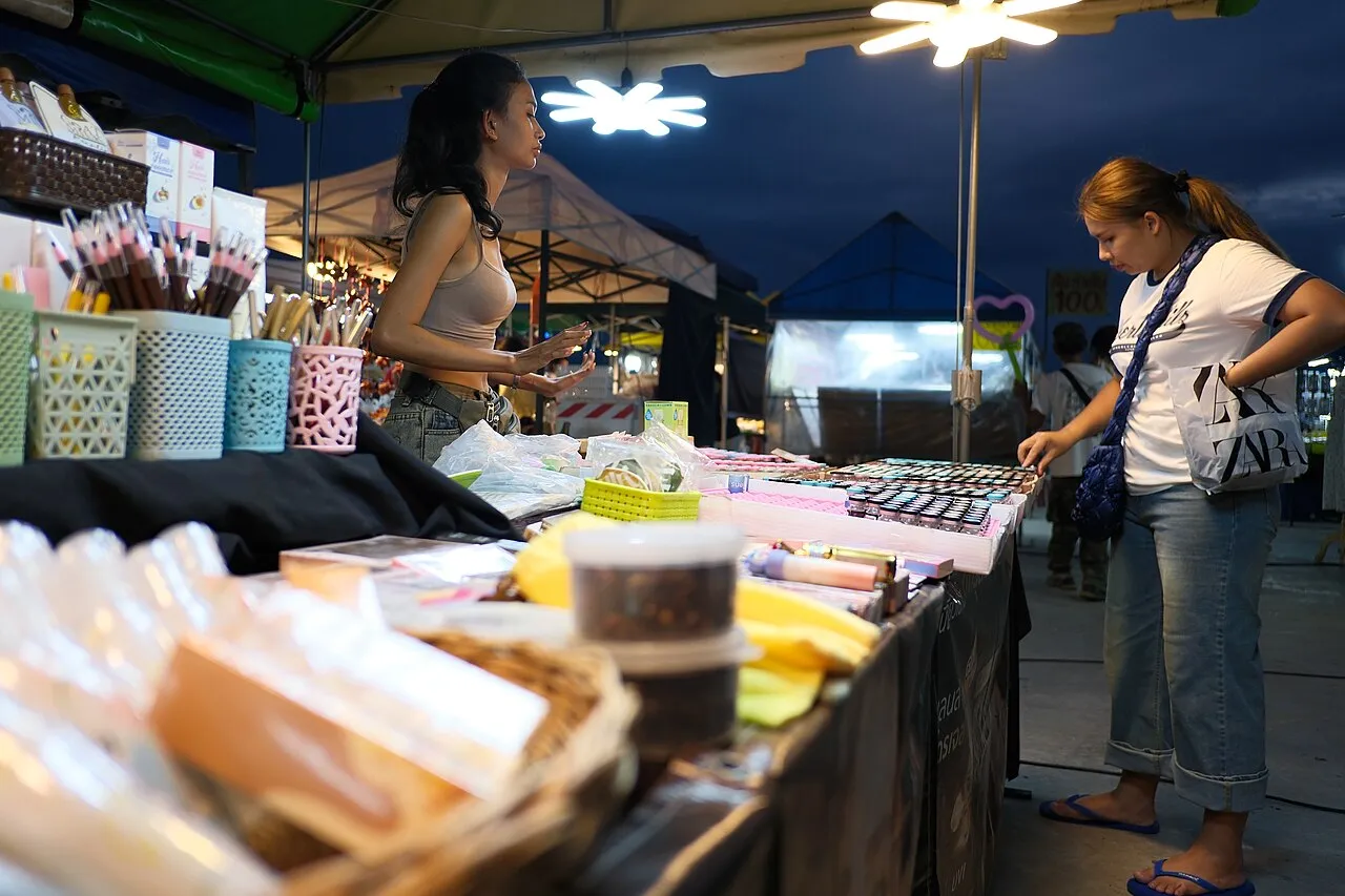 DSCF 0909 Two women browse and chat at a brightly lit outdoor market stall displaying an array of small goods and cosmetics at dusk